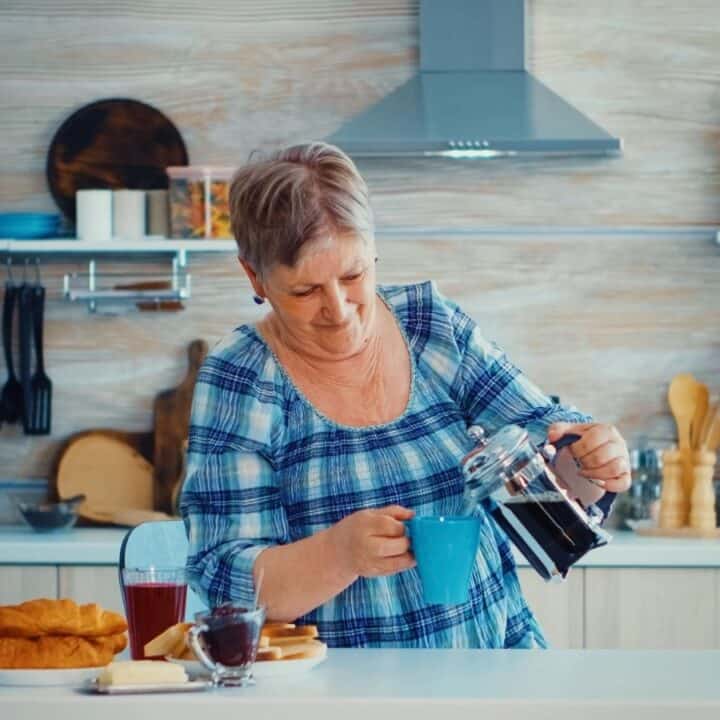 Older woman in a plaid shirt pours coffee from a French press into a blue mug in a modern kitchen with pastries and fruit on the counter.