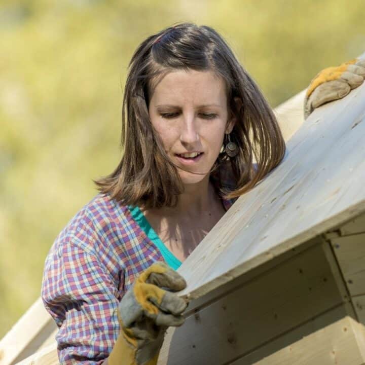 A person wearing gloves and a plaid shirt works on assembling or inspecting the wooden roof of an outdoor structure.