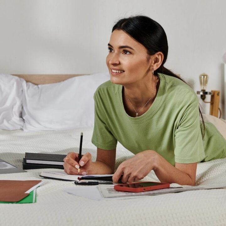 A woman lies on a bed writing in a notebook, with a laptop, books, and folders spread out, appearing to study or work.
