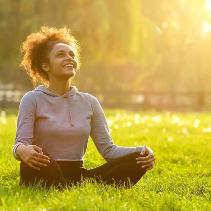A person sits cross-legged on grass in a park, smiling and looking up, surrounded by sunlight and greenery.
