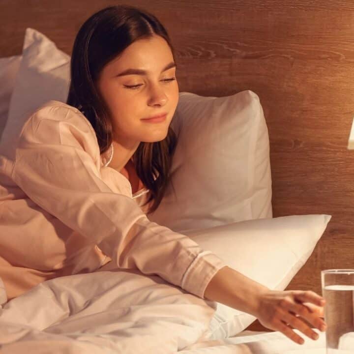 A woman in pajamas lies in bed and reaches for a glass of water on a nightstand next to a lit lamp.