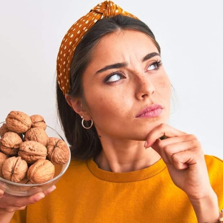A woman wearing a yellow top and headband holds a bowl of walnuts and looks to the side with a thoughtful expression.