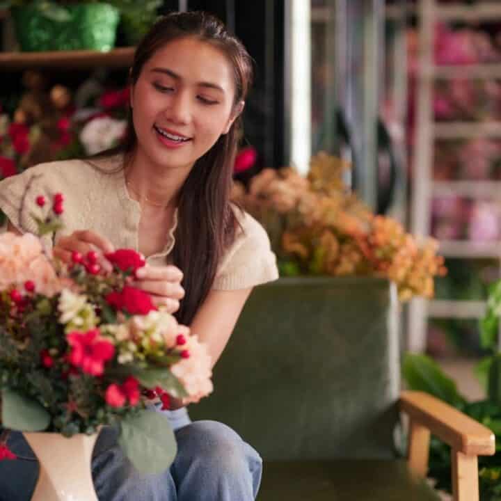 A woman sitting on a bench arranges flowers in a vase, surrounded by various plants and floral arrangements in a shop.