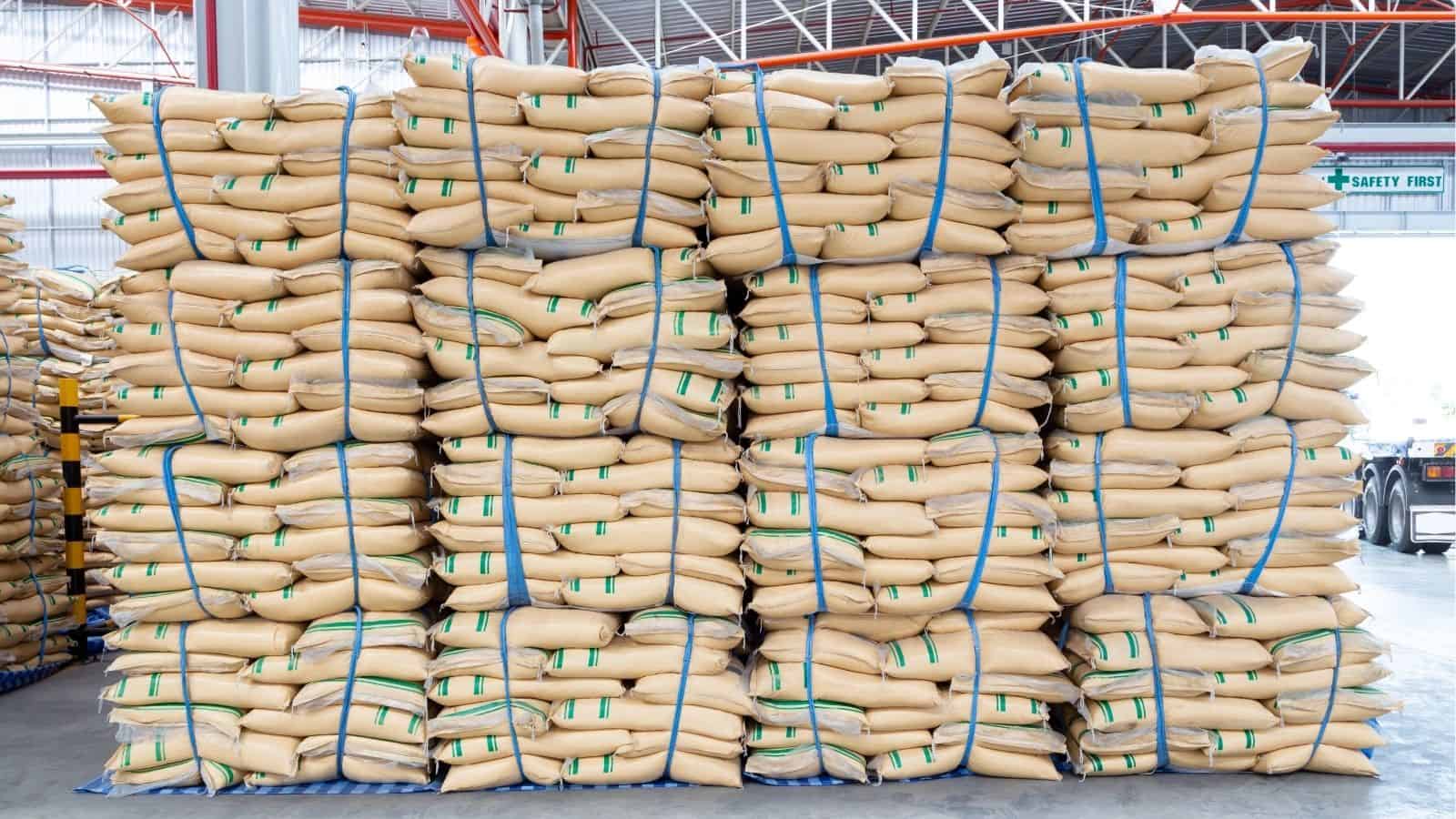 Stacks of large beige sacks, tightly bundled and arranged on pallets, are stored in a warehouse with a high ceiling and visible metal beams.