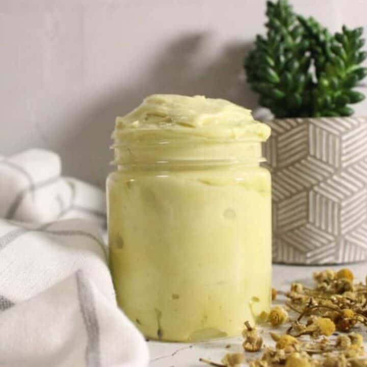 A glass jar filled with a light yellow cream sits on a white surface near dried chamomile flowers, a folded cloth, and a potted succulent.