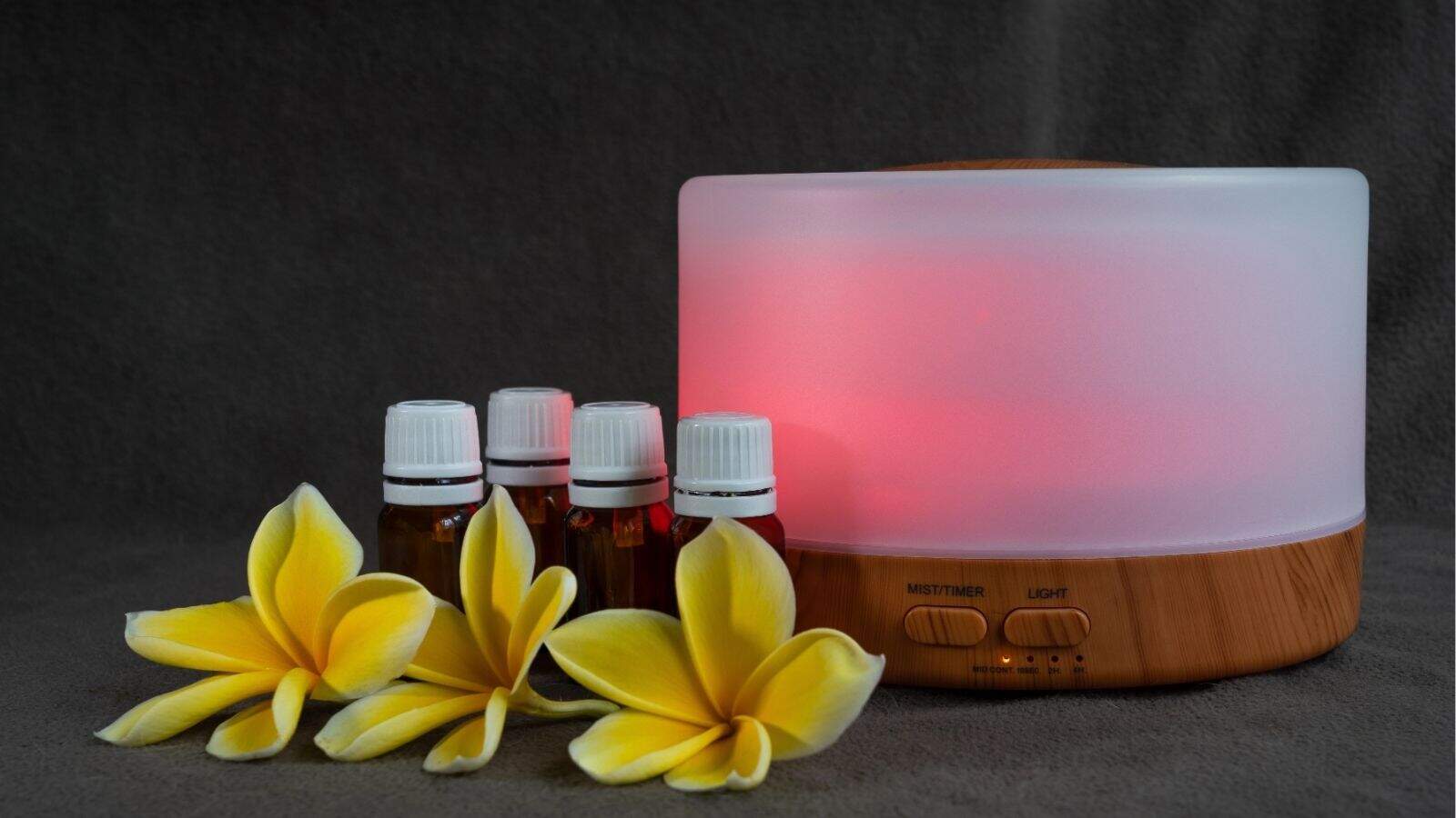 Aromatic diffuser with a glowing light next to five small essential oil bottles and three yellow plumeria flowers on a gray surface.