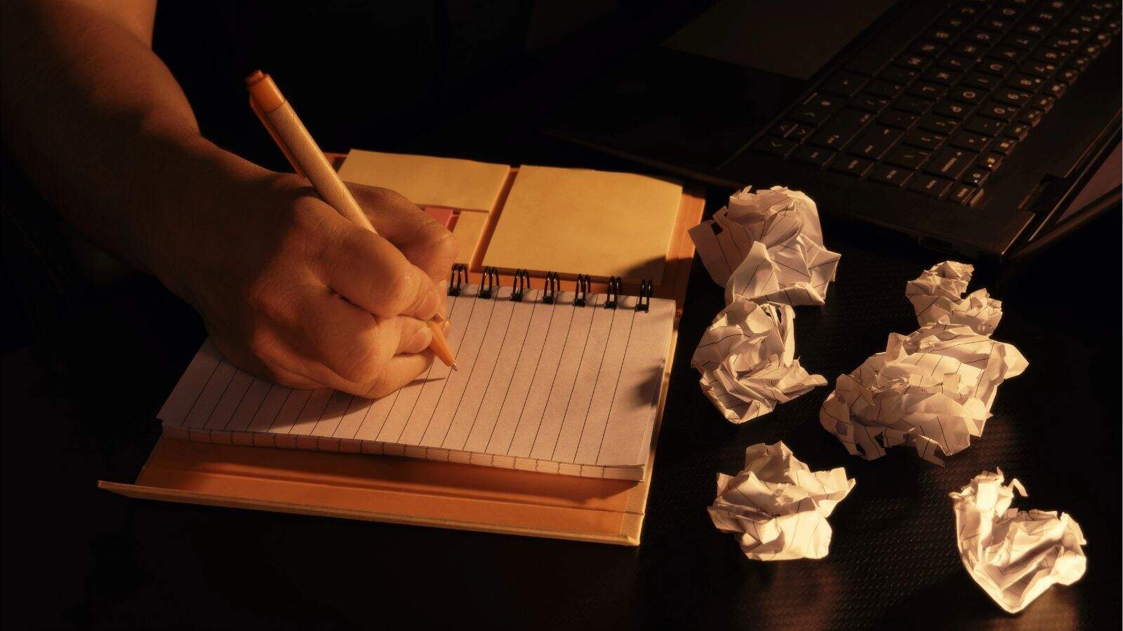 A person writes in a notebook beside a laptop, with several crumpled pieces of paper scattered on the desk.