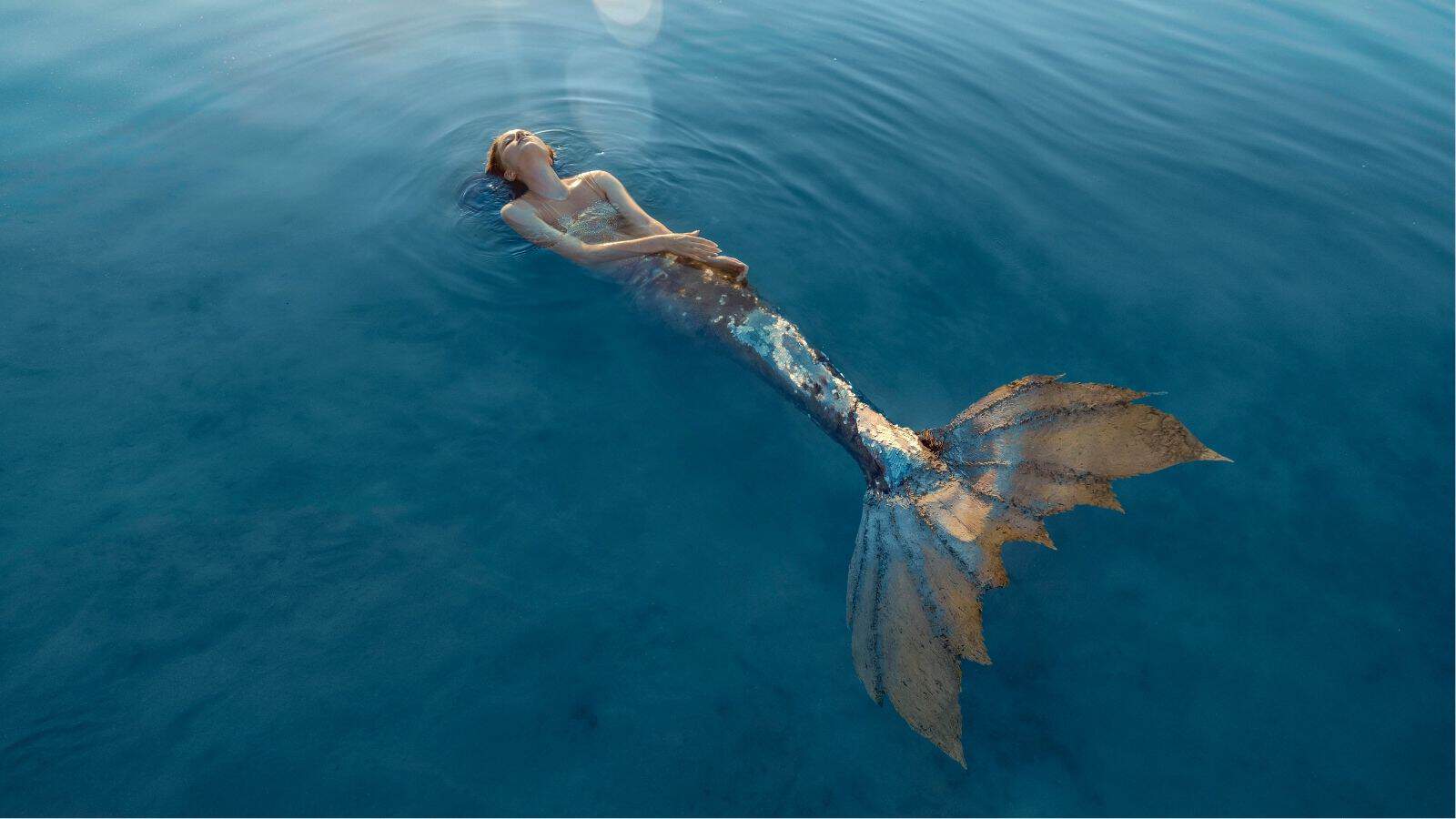 A person dressed as a mermaid floats on their back in calm, clear blue water.