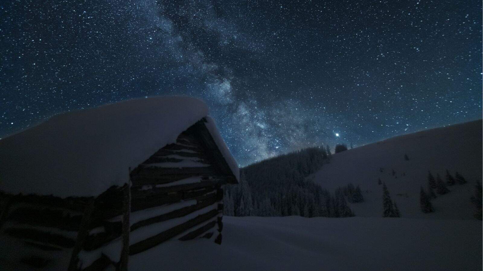 A snow-covered cabin sits on a snowy hillside under a clear night sky filled with stars and the Milky Way.