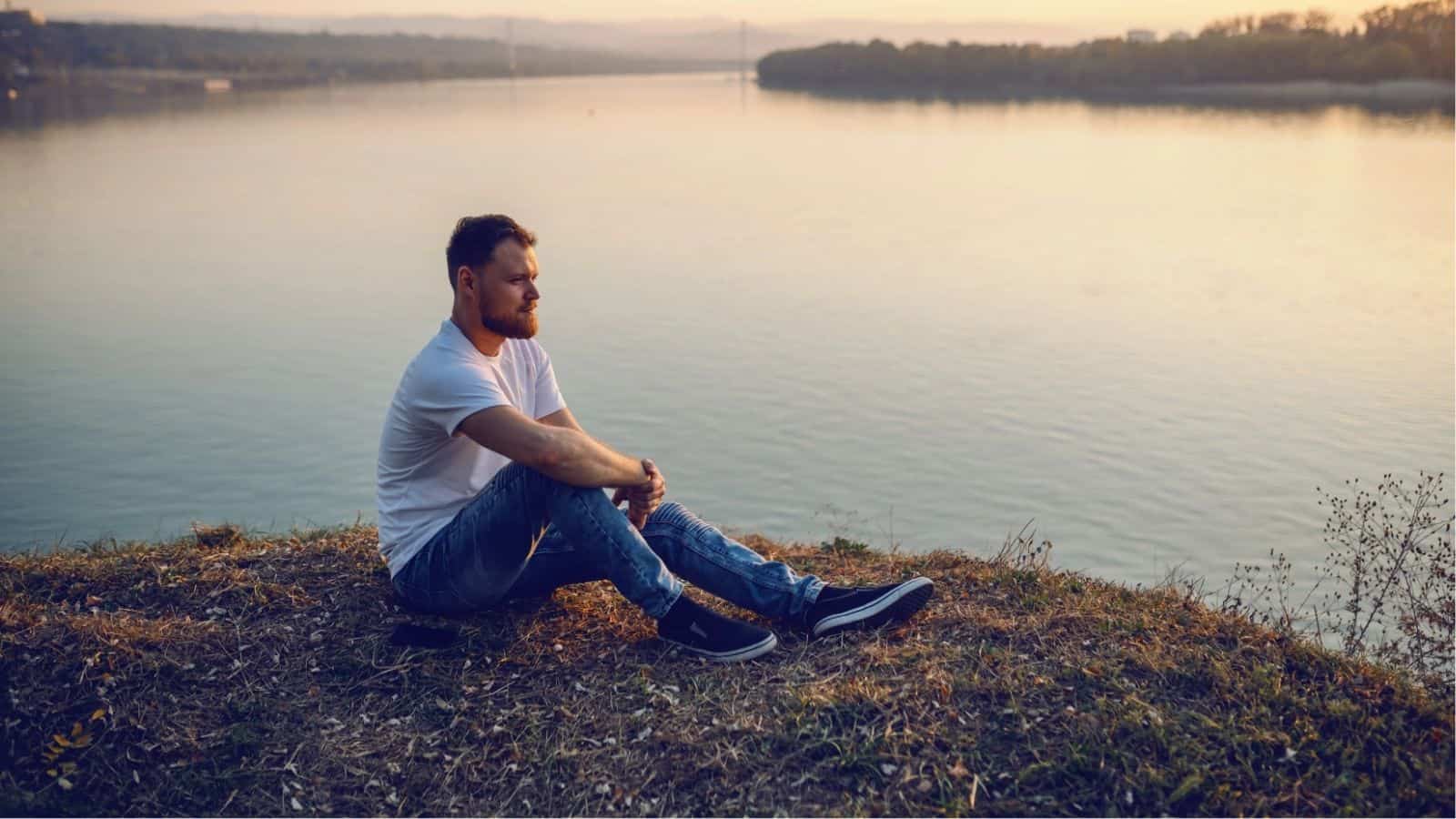 A man in a white t-shirt and jeans sits on the grass by a riverbank at sunset, looking out over the water.