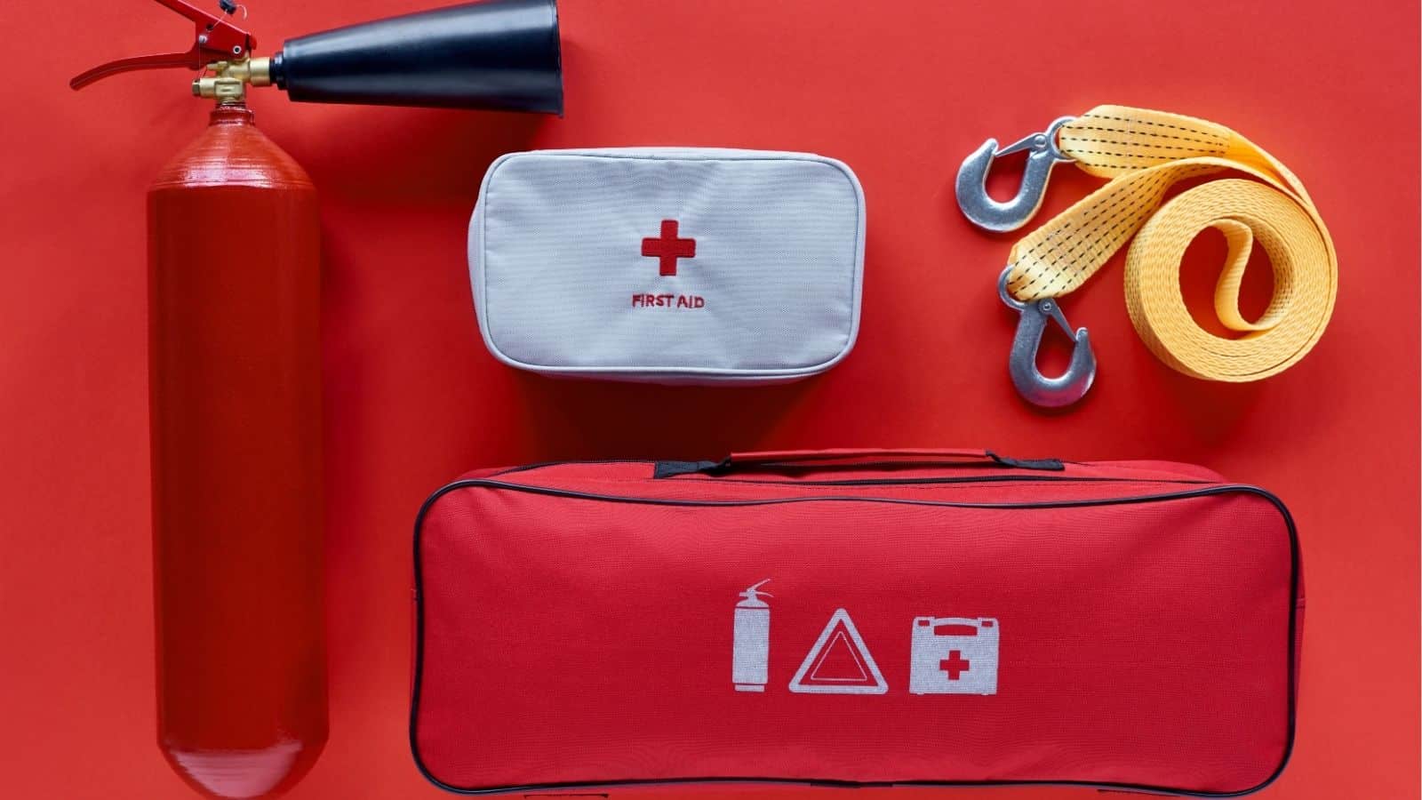 A fire extinguisher, a first aid kit, a red emergency kit bag, and a yellow tow strap with metal hooks arranged on a red surface.