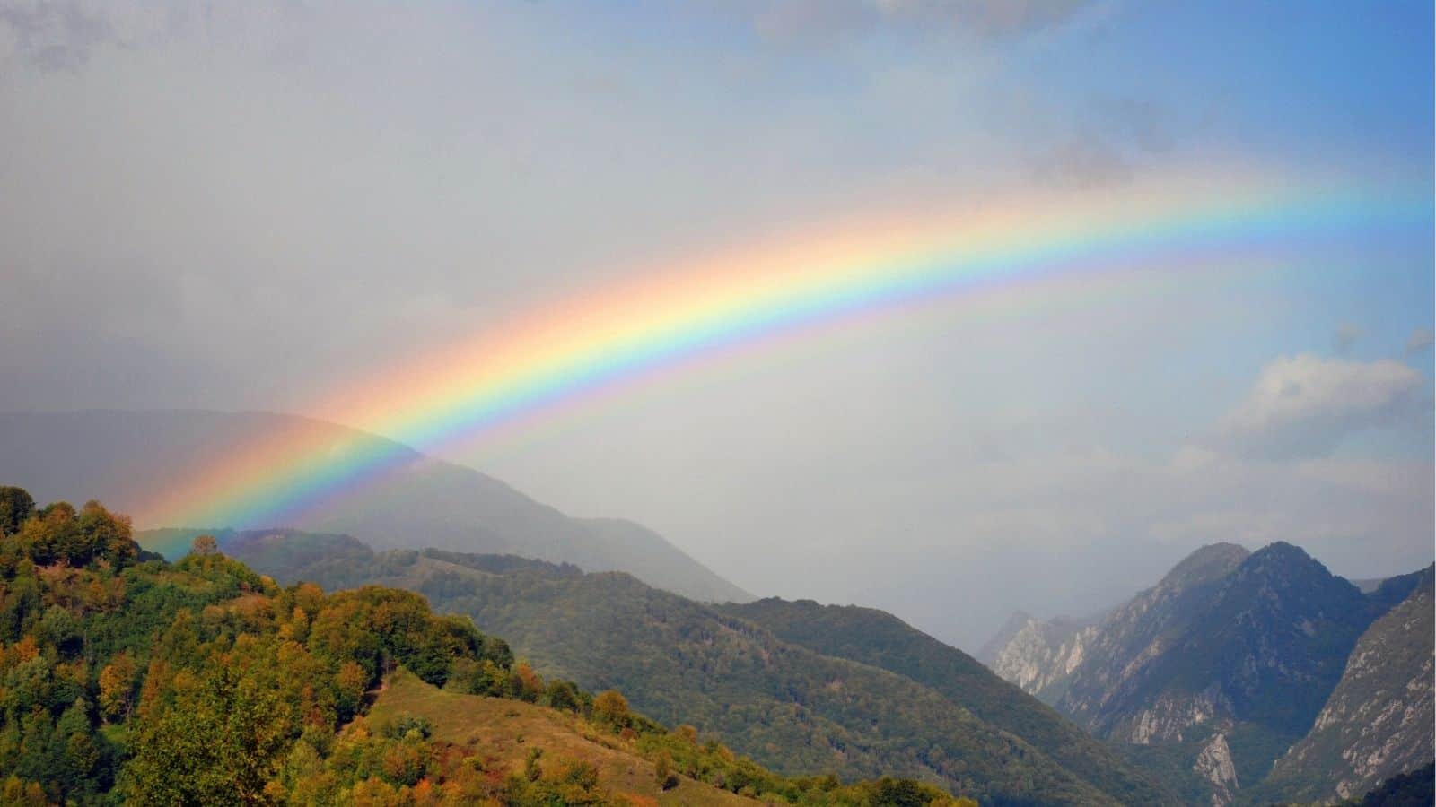 A bright rainbow arcs over green, forested hills and distant mountains under a partly cloudy sky.