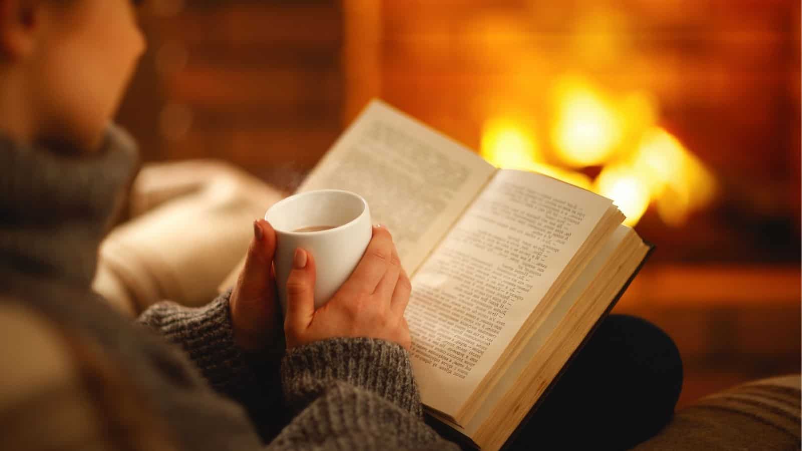 A person wearing a gray sweater holds a white mug and reads a book while sitting in front of a lit fireplace.