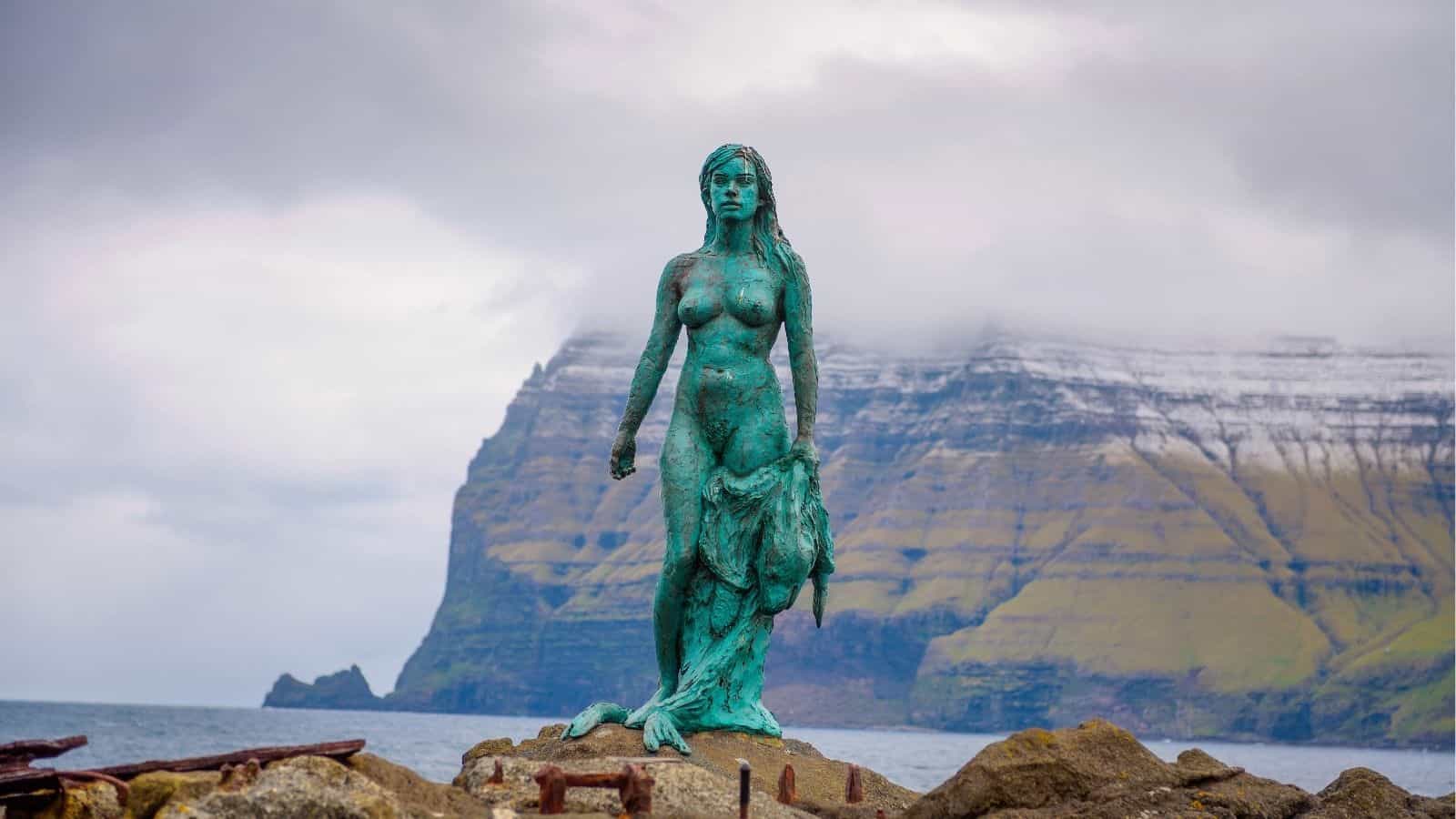 A bronze mermaid statue stands on rocks by the sea, with coastal cliffs and cloudy skies in the background.