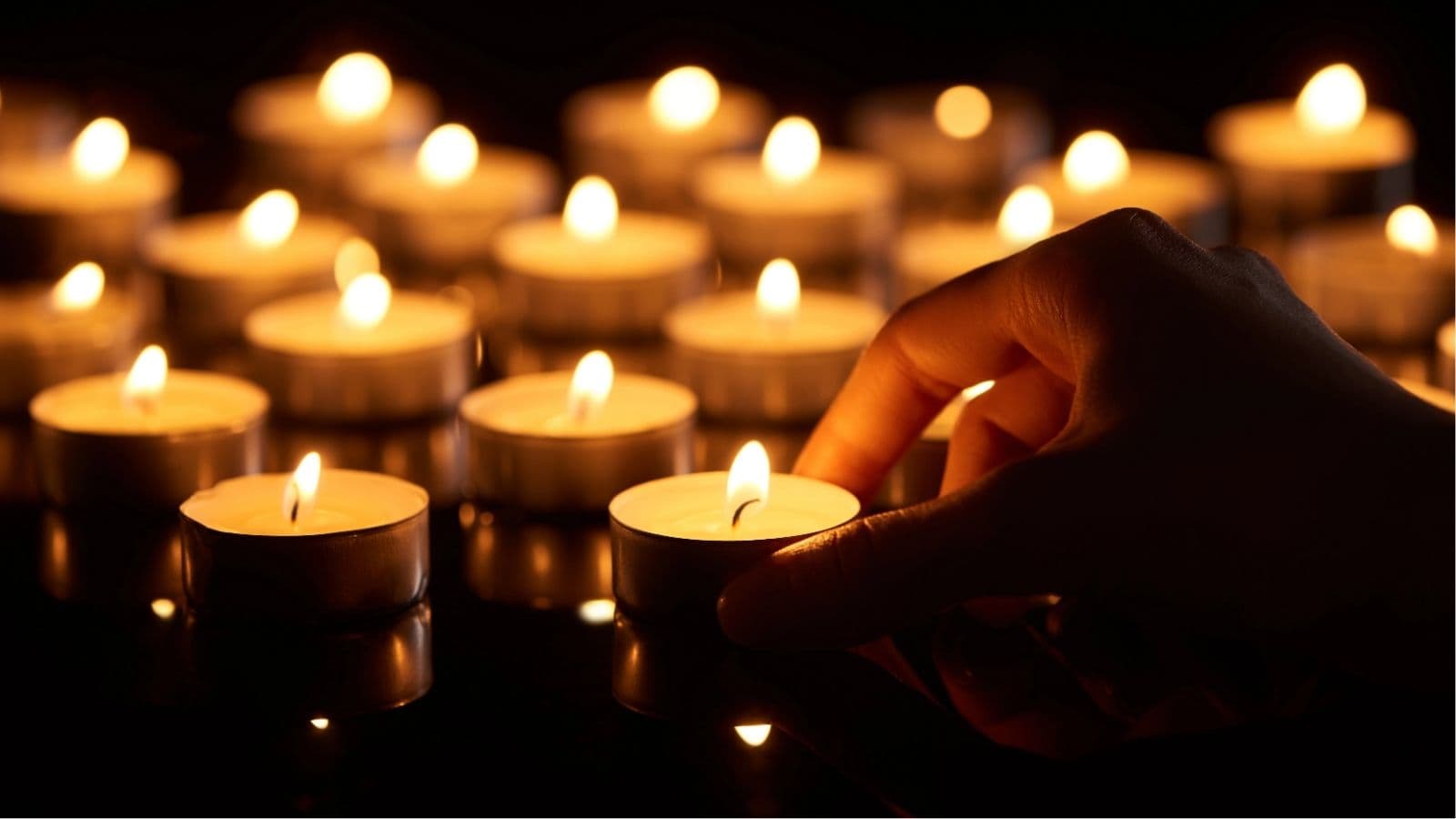 A hand reaches for a small lit tealight candle among several other glowing candles on a dark surface.