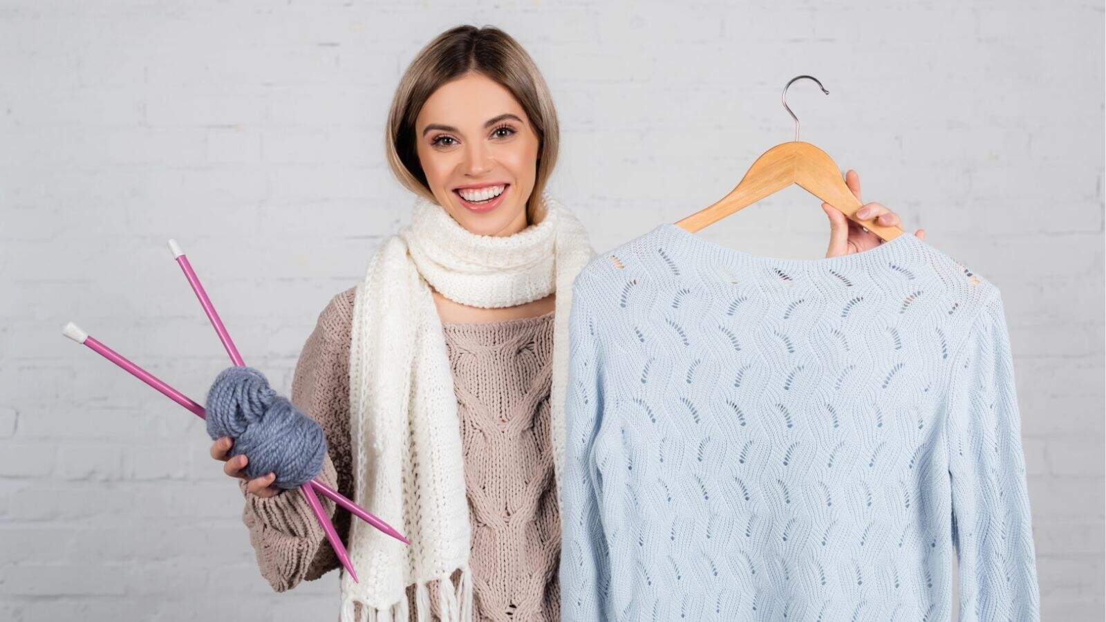 A woman stands smiling, holding knitting needles and yarn in one hand and a light blue knitted sweater on a hanger in the other.