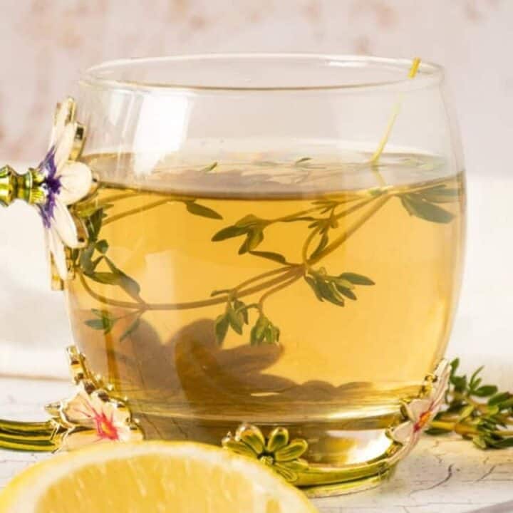 A glass cup with decorative floral handle filled with herbal tea, garnished with fresh thyme; a lemon wedge is in the foreground.