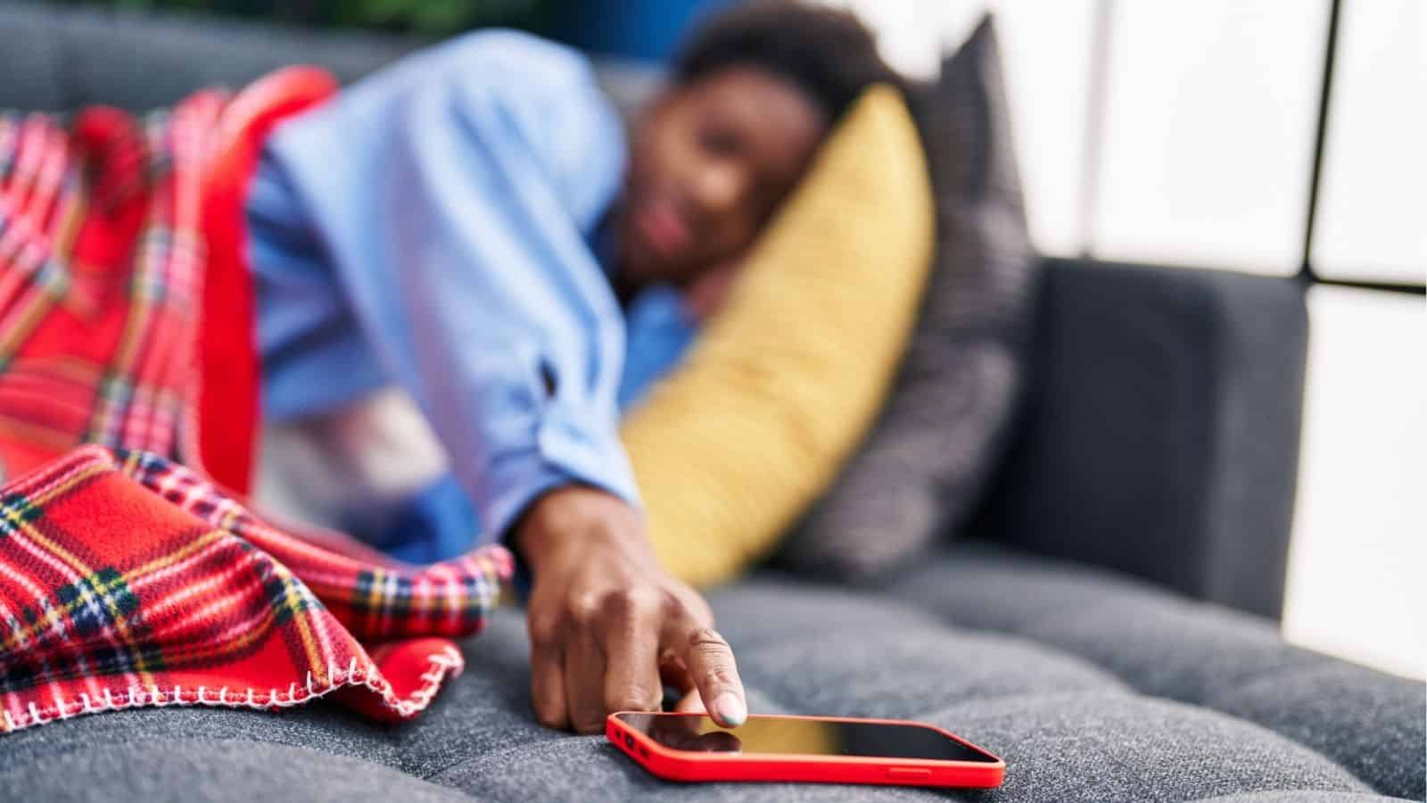 Person lying on a couch under a red plaid blanket, reaching out to touch a red smartphone on the couch.