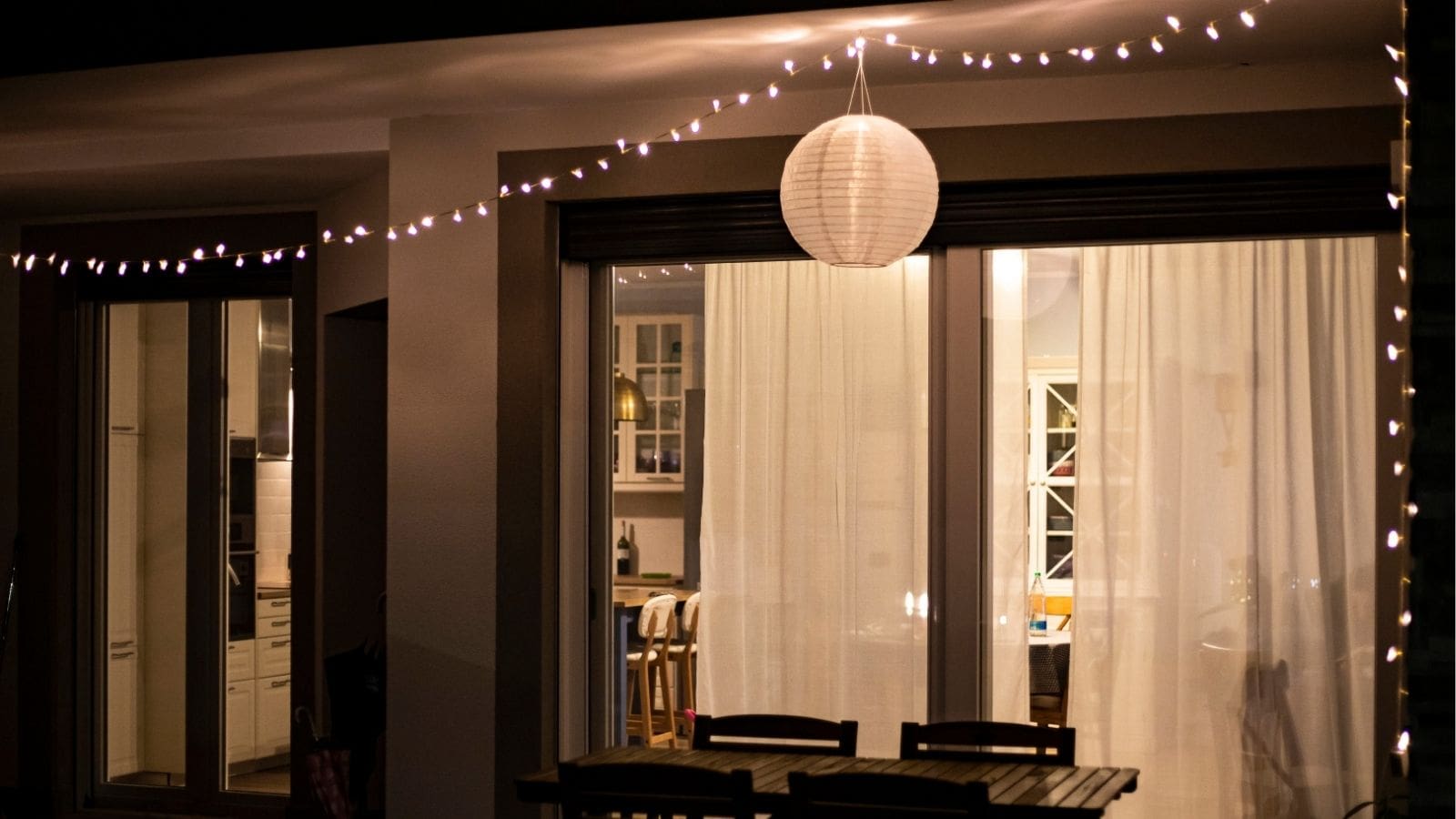 A patio with a wooden table and chairs at night, illuminated by string lights and a paper lantern. Large glass doors with curtains reveal a lit interior room.
