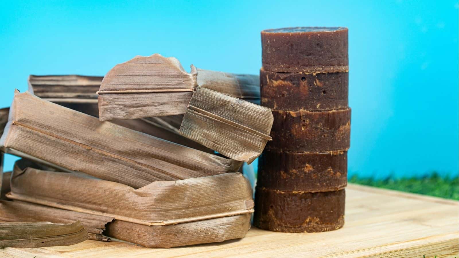 Brown blocks of palm sugar stacked beside wrapped pieces of unwrapped palm sugar on a wooden surface with a blue background.