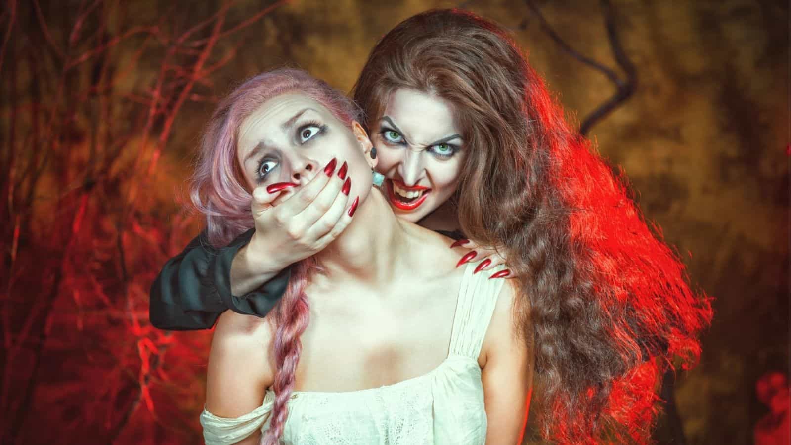 Two women in costume pose as a vampire attacking a victim; one woman bares fangs and grabs the other’s face amid a dark, eerie background with red lighting.