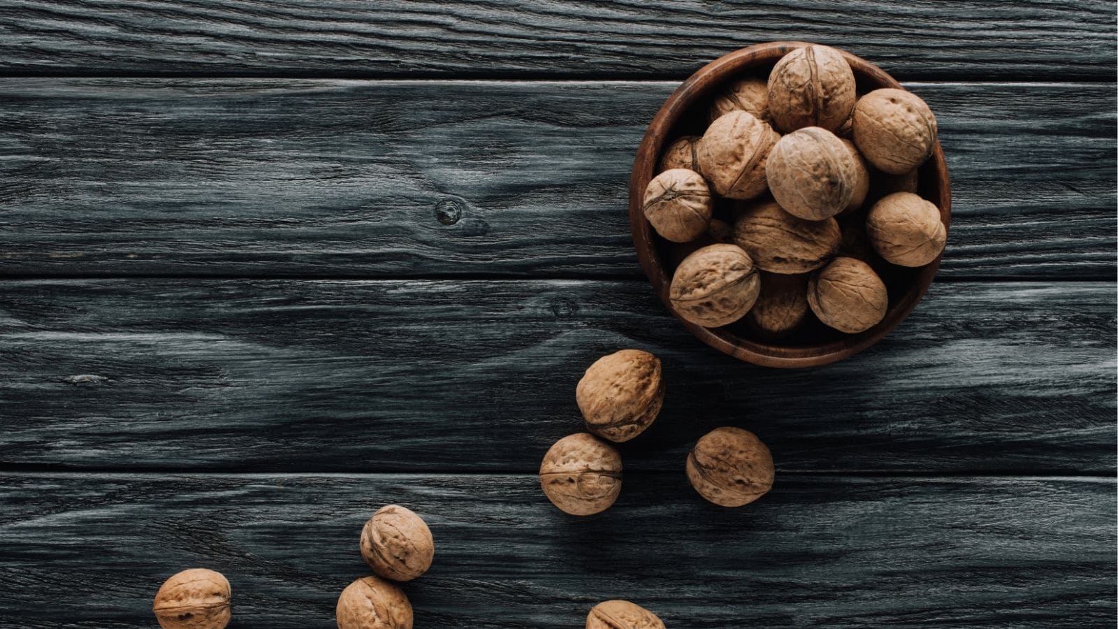 A wooden bowl filled with whole walnuts sits on a dark wooden surface, with several walnuts scattered nearby.