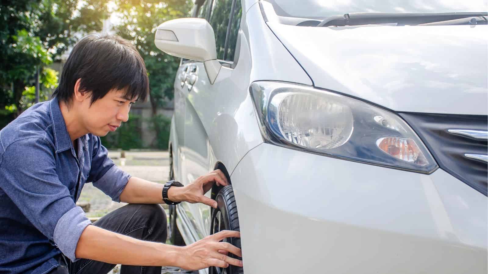 A person crouches beside a white car, inspecting or touching the front tire outdoors during daytime.
