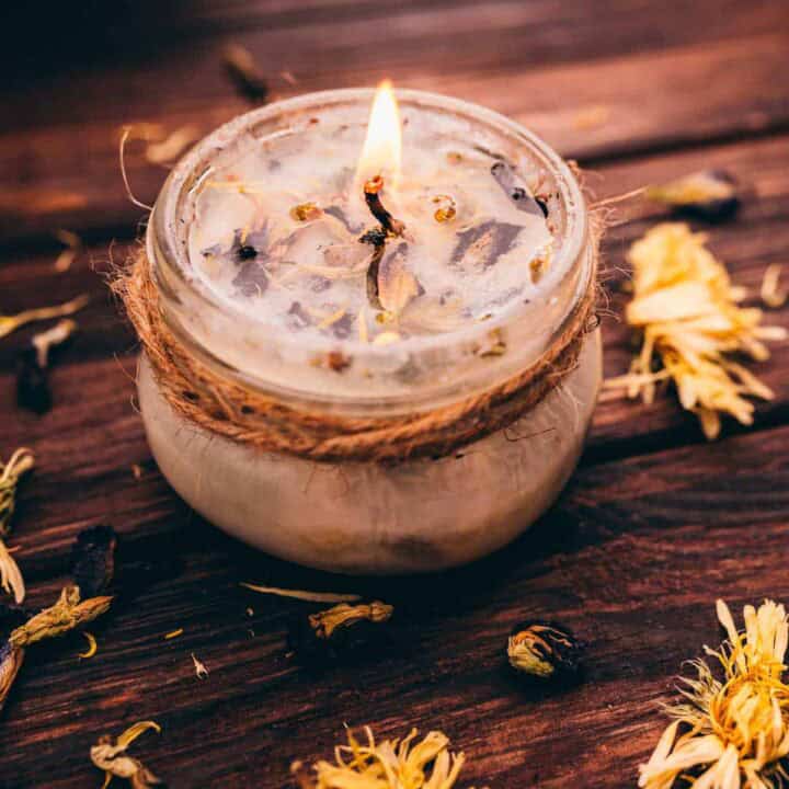 A small glass jar candle with dried flowers and herbs on top, burning on a wooden surface with scattered dried flowers around it.
