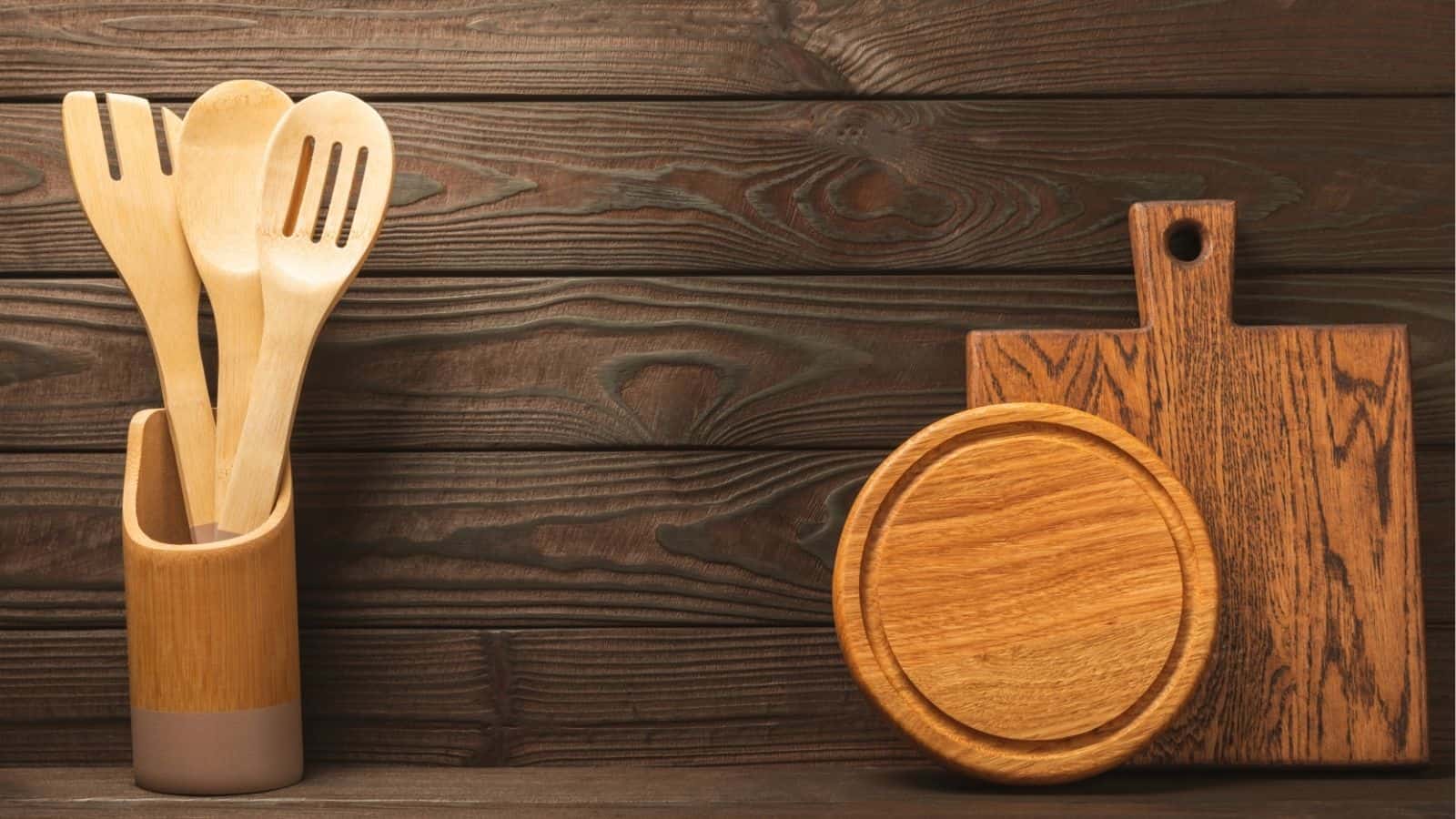 Wooden cooking utensils in a holder next to a round wooden plate and a rectangular cutting board on a dark wooden surface.