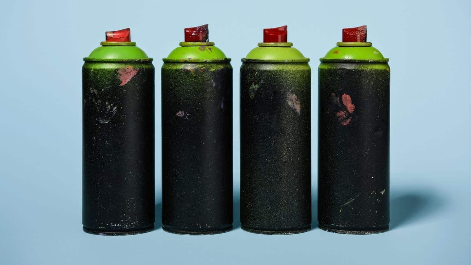 Four black spray paint cans with green tops and red nozzles are arranged in a row against a light blue background.