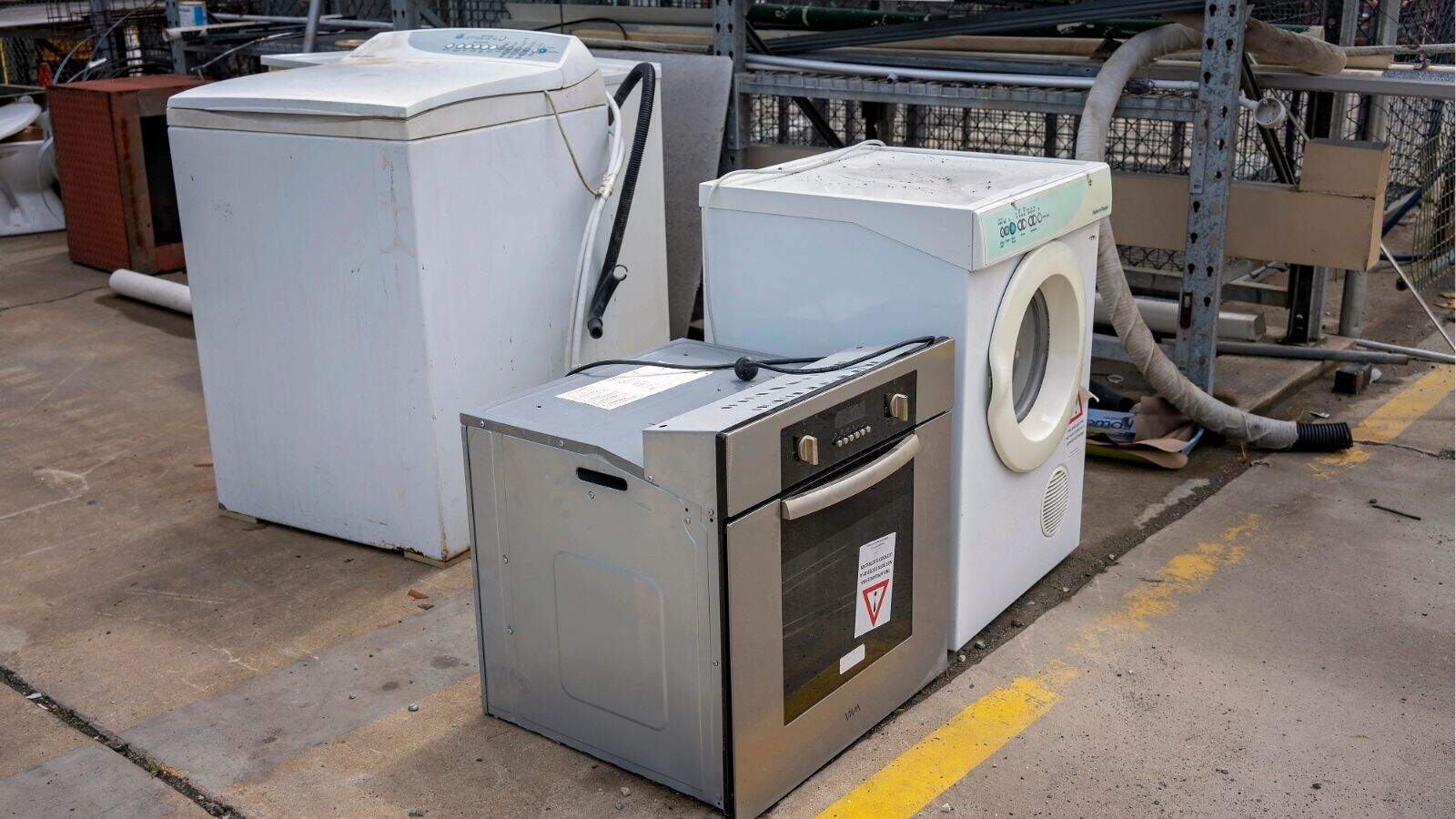 Three discarded household appliancesโa washing machine, a dryer, and an ovenโare placed on a concrete surface near a fence and other miscellaneous items.