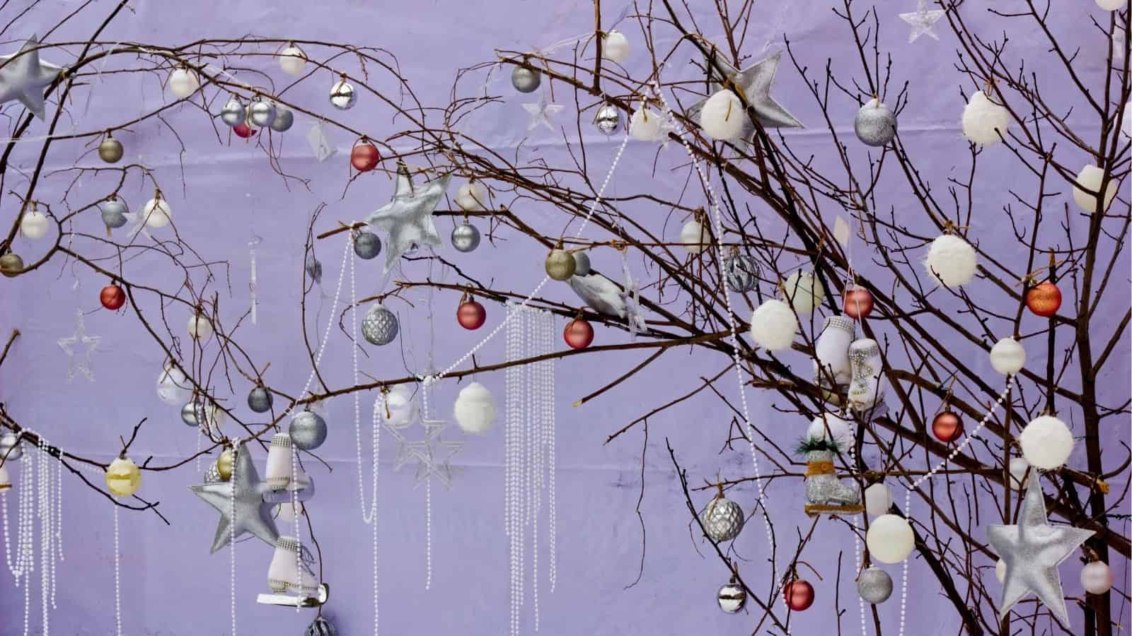 Bare tree branches decorated with silver, gold, and red ornaments, stars, and hanging beads against a purple wall.
