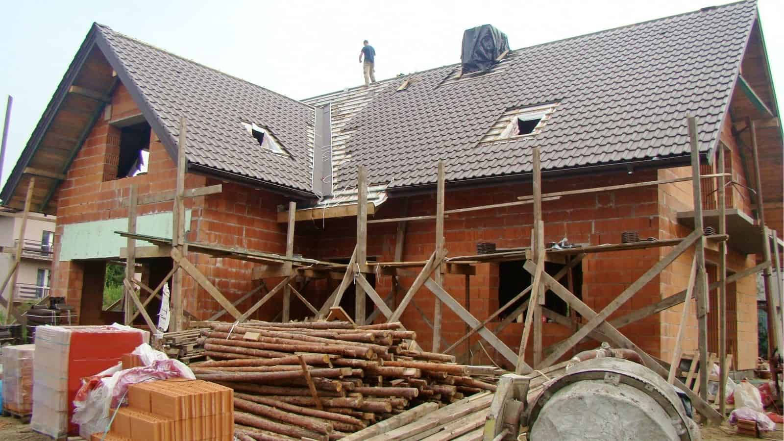 A two-story house under construction with scaffolding and building materials in front, and a worker standing on the roof.