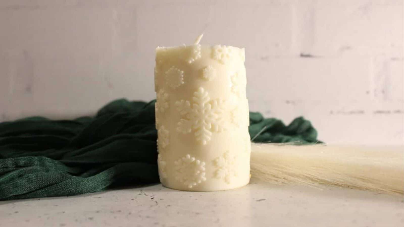 A white pillar candle with snowflake patterns sits on a white surface, with a dark green cloth and a light brush next to it against a white brick background.