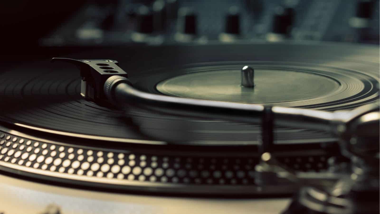Close-up of a vinyl record on a turntable with the tonearm and stylus positioned above the spinning record, with audio equipment blurred in the background.
