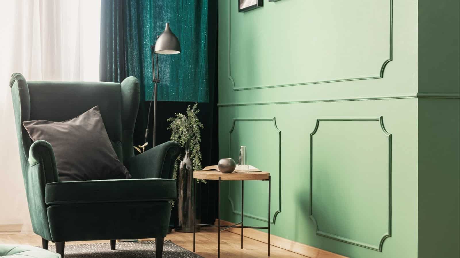 A green armchair with a black cushion sits beside a small round side table and floor lamp in a room with green paneled walls and a green curtain.