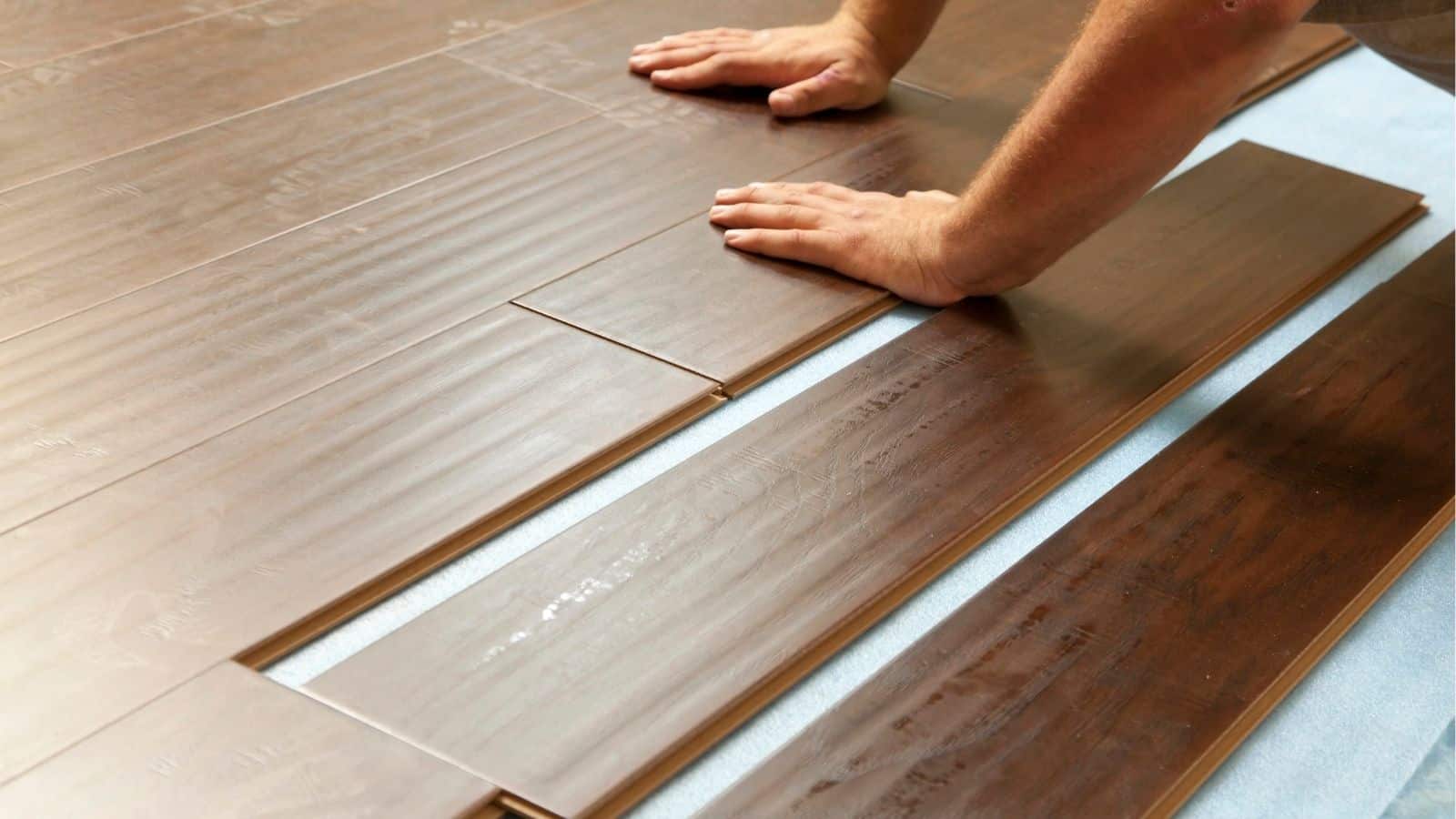 A person installs wood laminate flooring, aligning brown planks on a light underlayment with their hands.