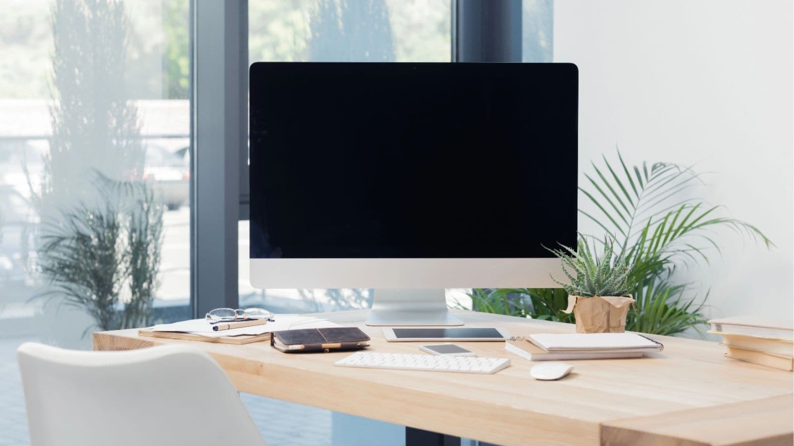 A desktop computer on a wooden desk with a keyboard, mouse, notepads, potted plants, and a smartphone in a modern, sunlit office space.