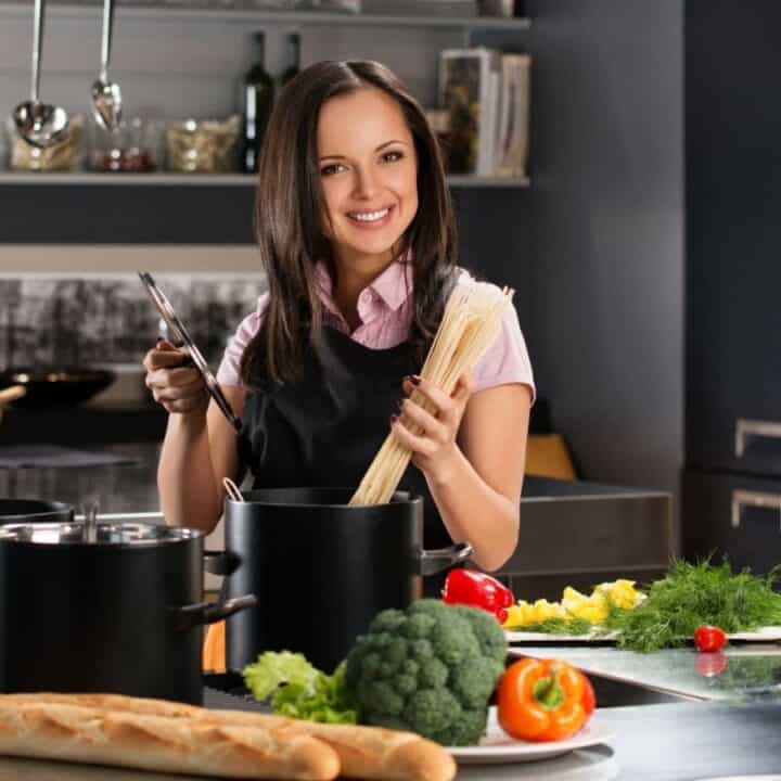 A woman stands in a modern kitchen preparing to cook pasta, surrounded by fresh vegetables, bread, and cooking utensils.