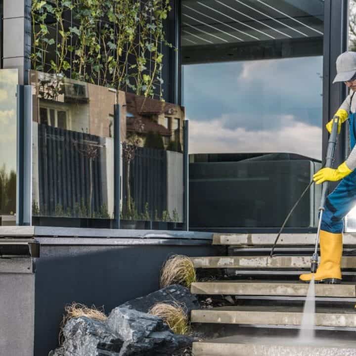 A person wearing yellow gloves and boots uses a pressure washer to clean outdoor steps next to a modern house with glass railings.