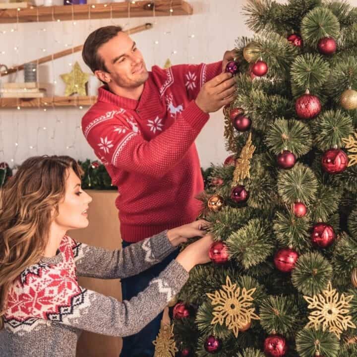 A man and a woman decorate a Christmas tree with ornaments in a living room decorated for the holidays.