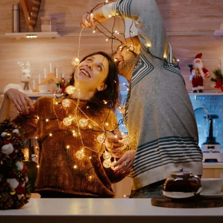 A woman and a man in a kitchen wrap themselves in string lights, smiling and decorating for Christmas. A Christmas tree and festive decorations are visible in the background.