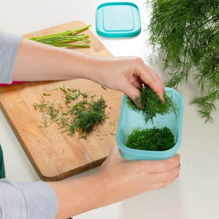 A person places freshly chopped dill into a blue container on a kitchen counter, with a cutting board and unused dill in the background.