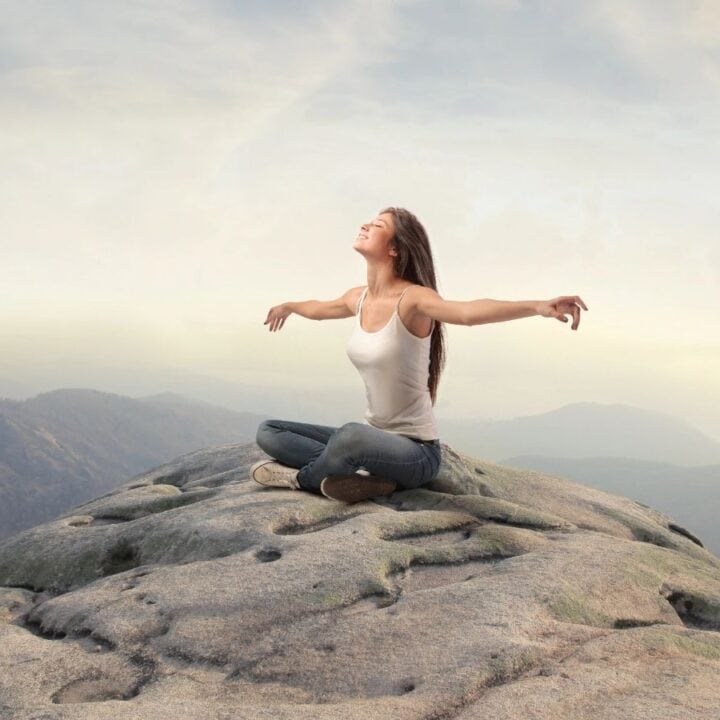 A woman sits cross-legged on a large rock at a mountain top with arms outstretched, facing the sky, with misty hills visible in the background.