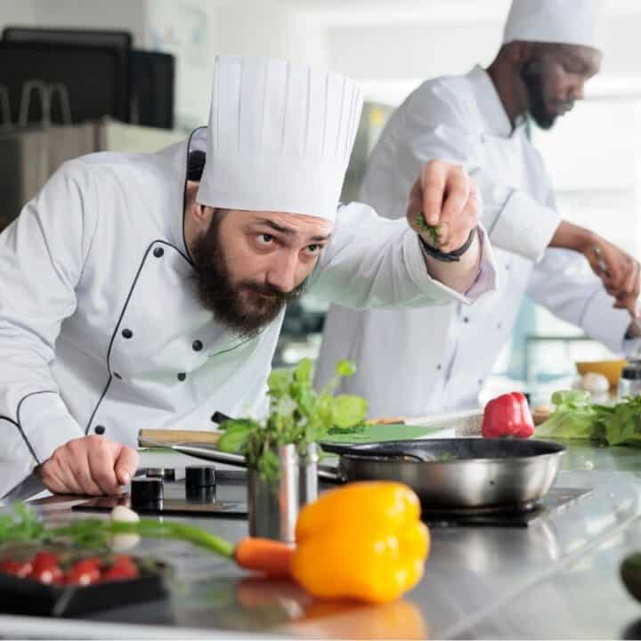 Two chefs in a kitchen, both wearing white uniforms and hats, prepare food; one garnishes a dish at the counter with various vegetables and ingredients.