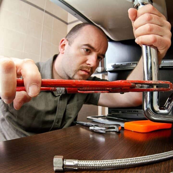 A man uses a red pipe wrench to tighten a metal pipe under a sink, with plumbing tools and parts visible on the countertop.