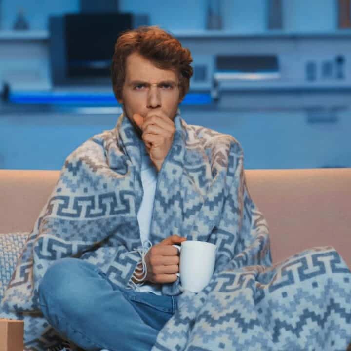 A man sits on a couch wrapped in a blanket, holding a white mug and coughing, with tissues nearby, suggesting he is unwell.