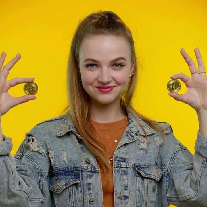 A young woman in a denim jacket holds a gold coin in each hand, standing against a bright yellow background.
