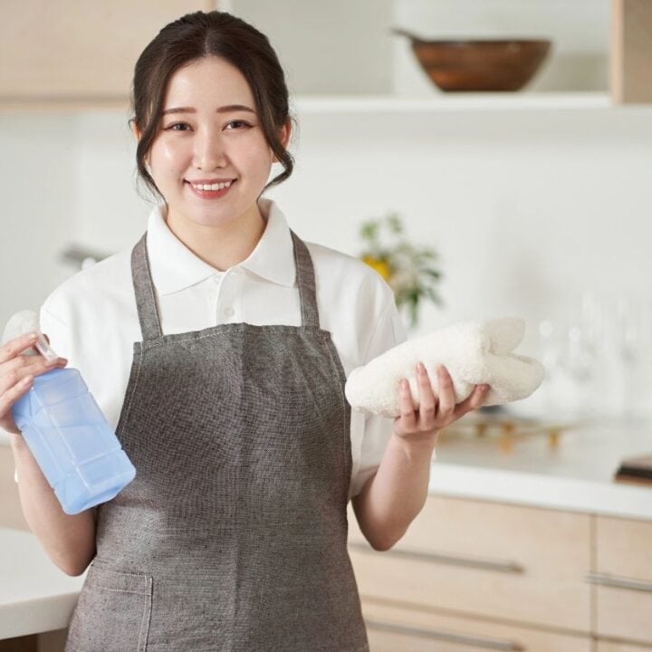 A woman in an apron stands in a kitchen holding a spray bottle in one hand and a folded towel in the other, smiling at the camera.