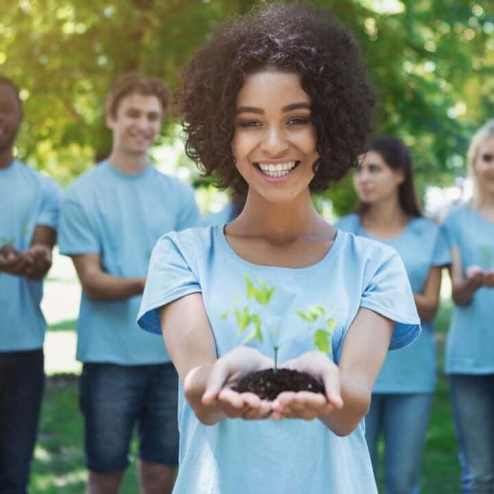 A group of people wearing light blue shirts stand outside. The woman in front holds soil with small green plants sprouting in her hands and smiles at the camera.